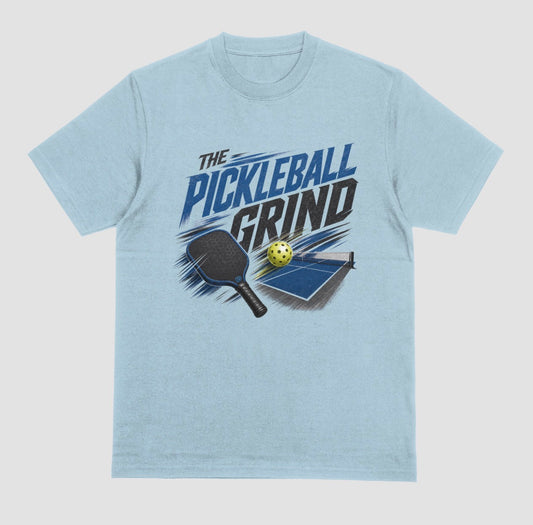 A light blue short-sleeve t-shirt with a graphic on the front that reads 'THE PICKLEBALL GRIND' in blue and black, accompanied by illustrations of a pickleball paddle, ball, and table.