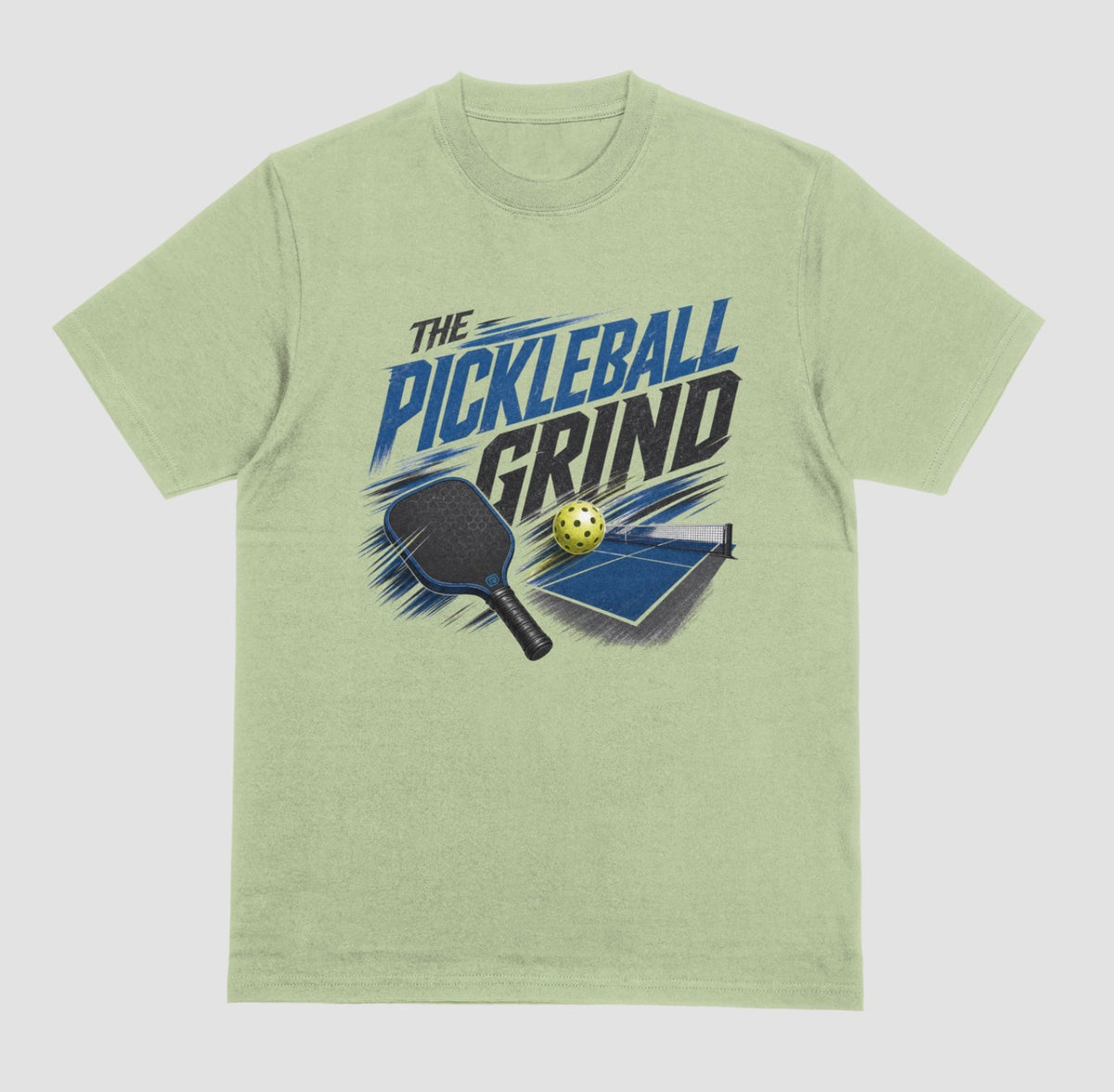 A pistachio green short-sleeve t-shirt with a graphic on the front that reads 'THE PICKLEBALL GRIND' in blue and black, accompanied by illustrations of a pickleball paddle, ball, and table.