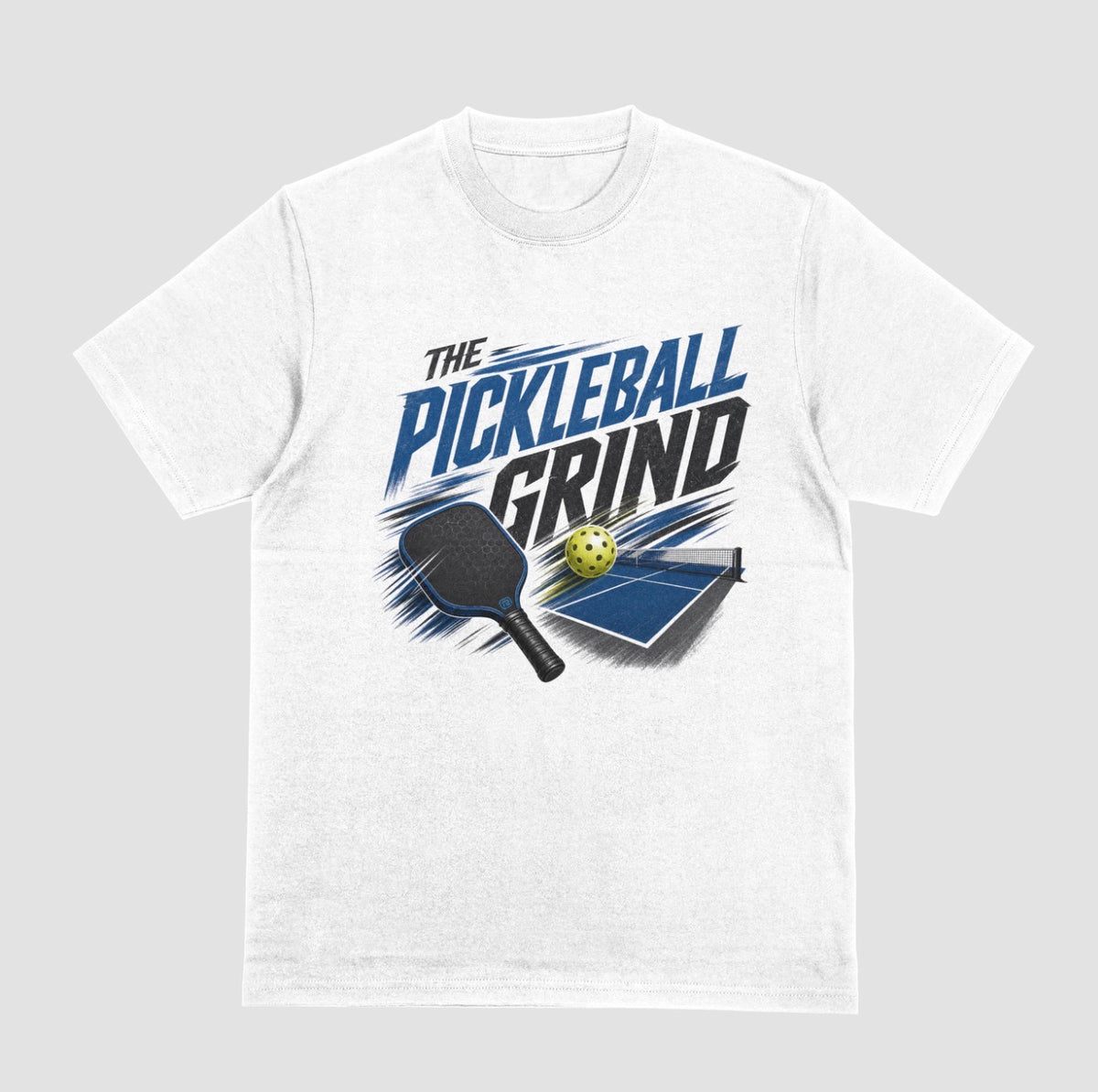 A white short-sleeve t-shirt with a graphic on the front that reads 'THE PICKLEBALL GRIND' in blue and black, accompanied by illustrations of a pickleball paddle, ball, and table.