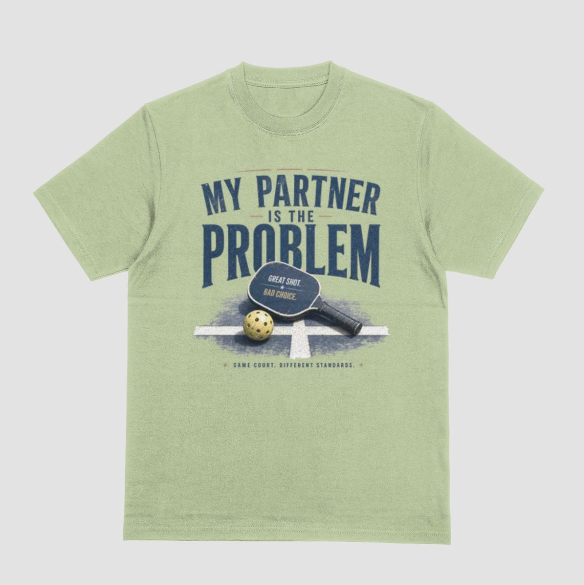 A pistachio green short-sleeve t-shirt with a vintage-style graphic on the front. The graphic features the text 'MY PARTNER IS THE PROBLEM' in dark blue, along with a paddle, ball, and net illustration.