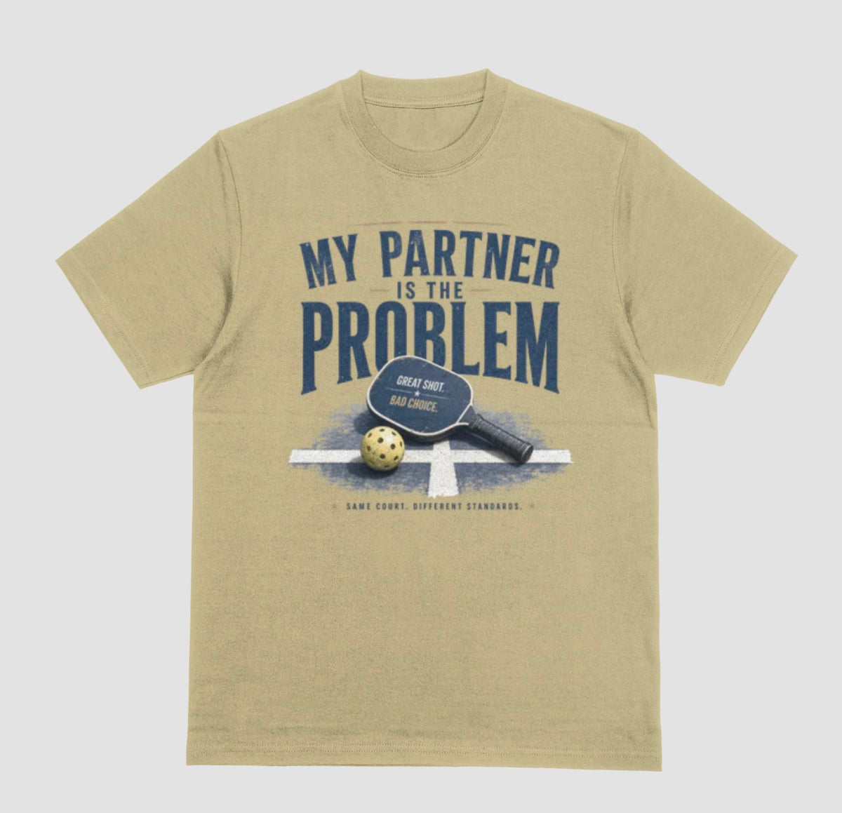 A sand color short-sleeve t-shirt with a vintage-style graphic on the front. The graphic features the text 'MY PARTNER IS THE PROBLEM' in dark blue, along with a paddle, ball, and net illustration.