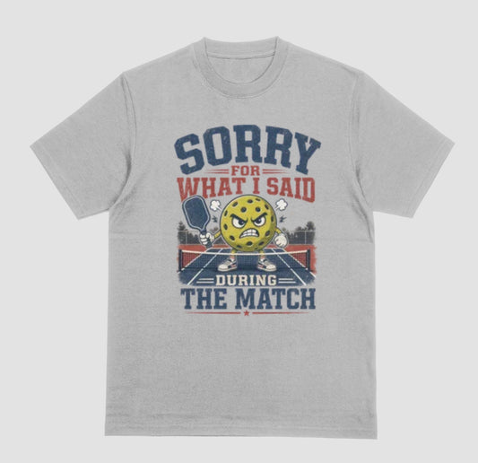 A gray short-sleeve t-shirt with a distressed graphic on the front. The graphic features a yellow cartoon pickleball with an angry face, a pickleball paddle, and the text 'SORRY FOR WHAT I SAID DURING THE MATCH' in navy blue and red lettering, set against a solid light gray background.