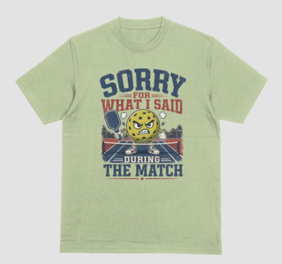 A pistachio green short-sleeve t-shirt with a distressed graphic on the front. The graphic features a yellow cartoon pickleball with an angry face, a pickleball paddle, and the text 'SORRY FOR WHAT I SAID DURING THE MATCH' in navy blue and red lettering, set against a solid light gray background.