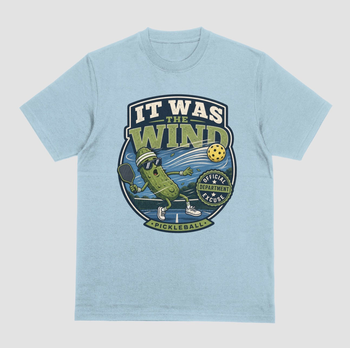 A light blue crewneck t-shirt featuring a vintage-style circular graphic on the front with a pickleball-themed cartoon character and the text 'IT WAS THE WIND' The Official Excuse Department.
