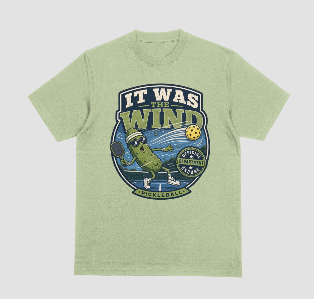 A pistachio green crewneck t-shirt featuring a vintage-style circular graphic on the front with a pickleball-themed cartoon character and the text 'IT WAS THE WIND' The Official Excuse Department.