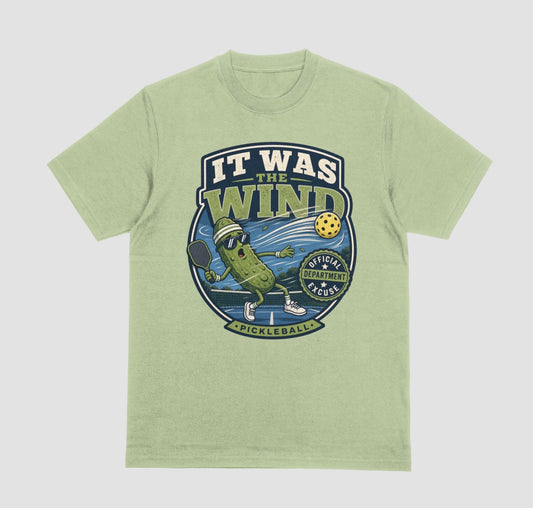 A pistachio green crewneck t-shirt featuring a vintage-style circular graphic on the front with a pickleball-themed cartoon character and the text 'IT WAS THE WIND' The Official Excuse Department.