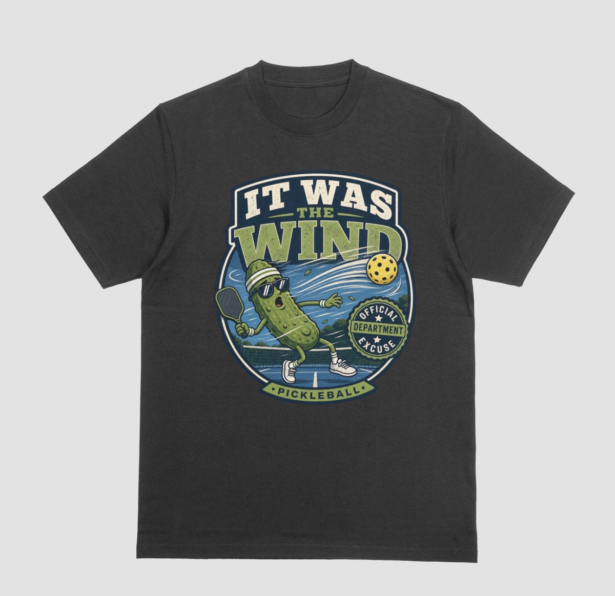 A black crewneck t-shirt featuring a vintage-style circular graphic on the front with a pickleball-themed cartoon character and the text 'IT WAS THE WIND' The Official Excuse Department.