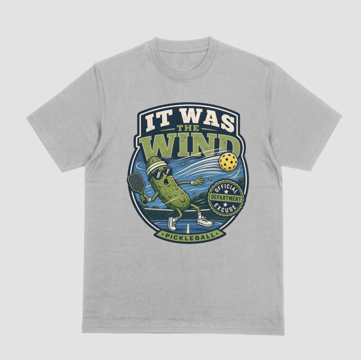 A gray crewneck t-shirt featuring a vintage-style circular graphic on the front with a pickleball-themed cartoon character and the text 'IT WAS THE WIND' The Official Excuse Department.