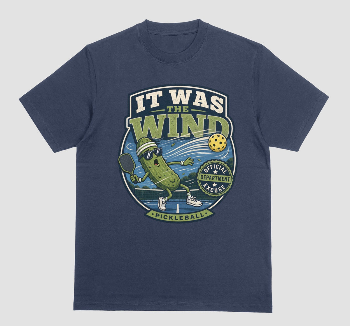 A navy blue crewneck t-shirt featuring a vintage-style circular graphic on the front with a pickleball-themed cartoon character and the text 'IT WAS THE WIND' The Official Excuse Department.