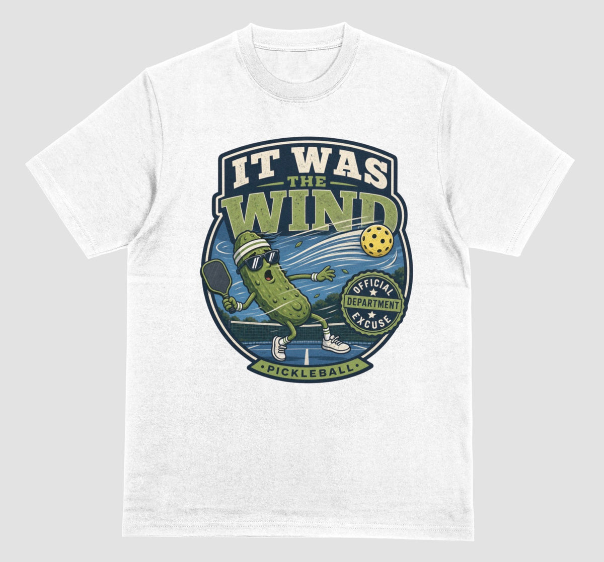 A white crewneck t-shirt featuring a vintage-style circular graphic on the front with a pickleball-themed cartoon character and the text 'IT WAS THE WIND' The Official Excuse Department.