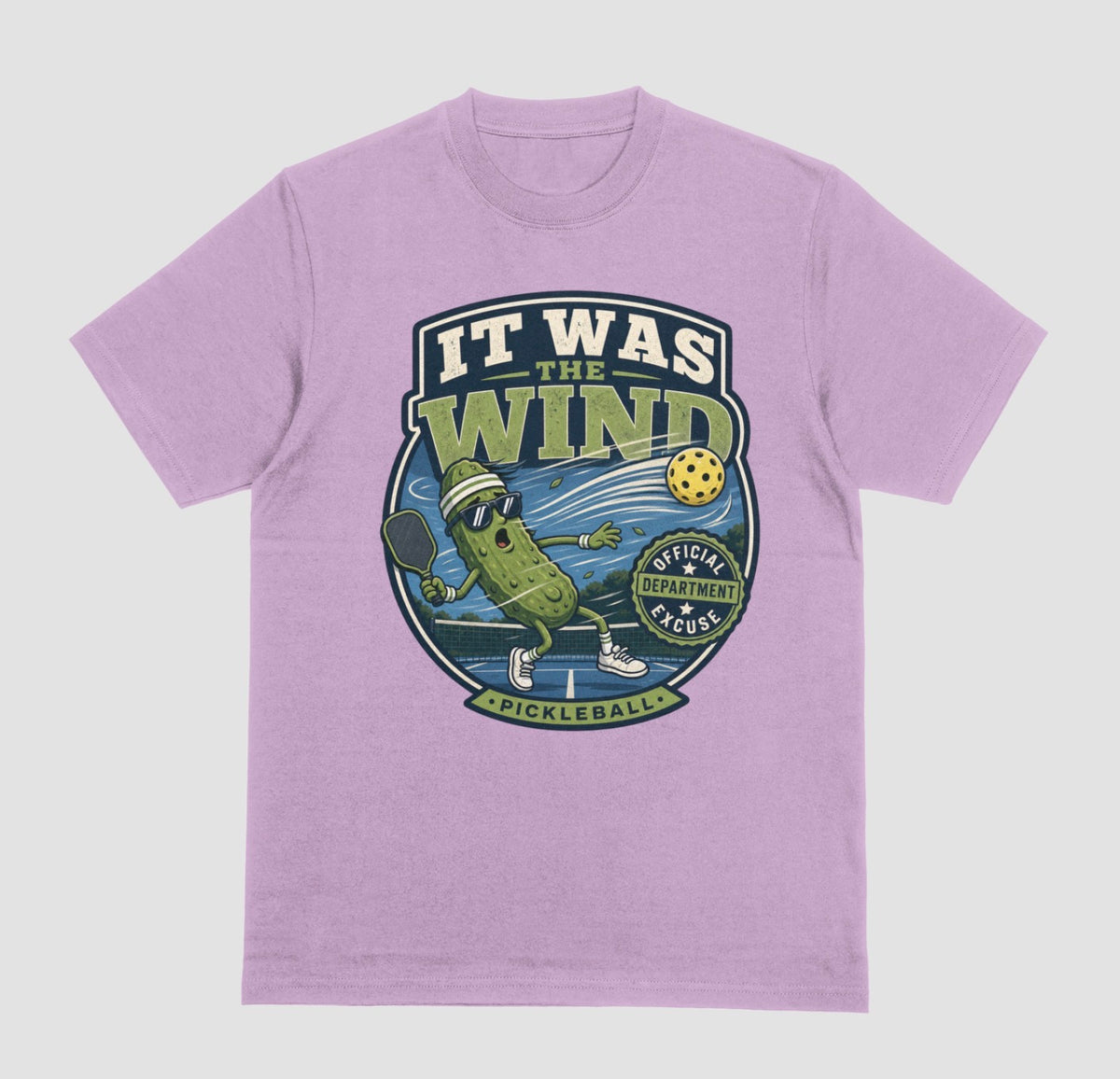 A lilac crewneck t-shirt featuring a vintage-style circular graphic on the front with a pickleball-themed cartoon character and the text 'IT WAS THE WIND' The Official Excuse Department.