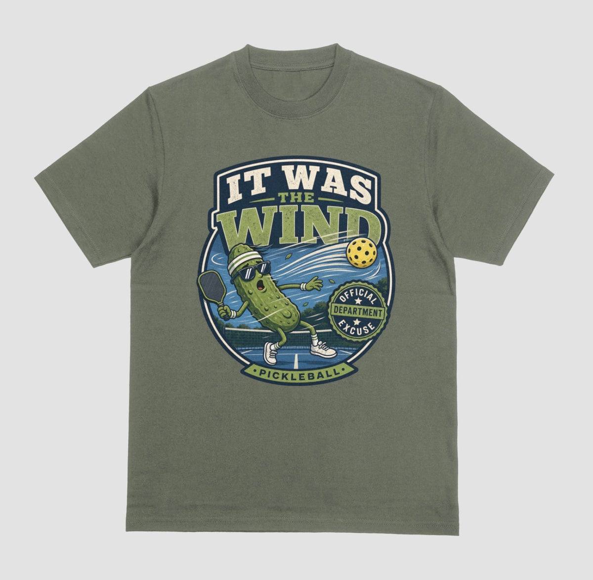A military green crewneck t-shirt featuring a vintage-style circular graphic on the front with a pickleball-themed cartoon character and the text 'IT WAS THE WIND' The Official Excuse Department.