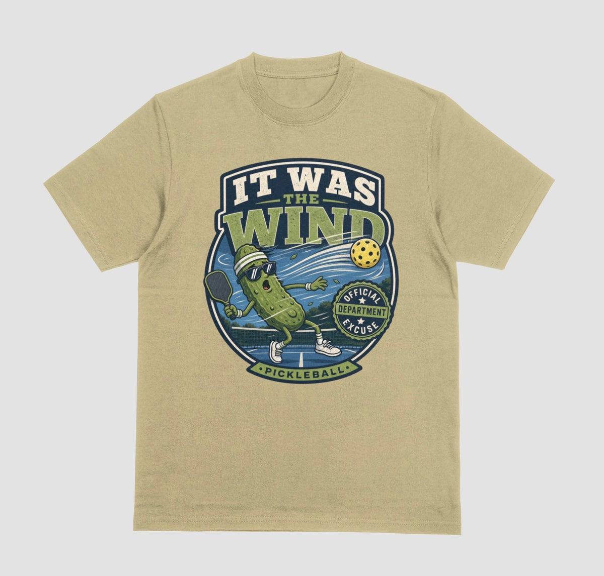 A sand color crewneck t-shirt featuring a vintage-style circular graphic on the front with a pickleball-themed cartoon character and the text 'IT WAS THE WIND' The Official Excuse Department.