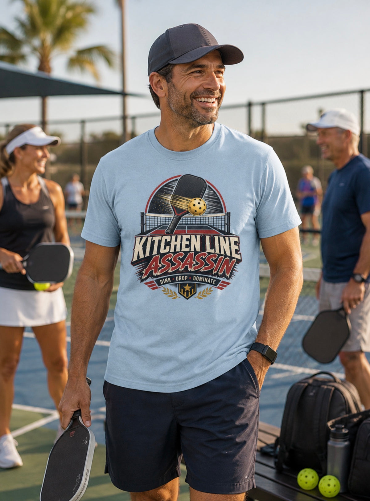 A man on an outdoor pickleball court wearing a light blue graphic t-shirt that reads 'KITCHEN LINE ASSASSIN' with a pickleball graphic, navy shorts, and a dark cap.