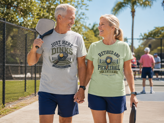 A couple walking on a pickleball court. The man wearing t-shirt with a vintage-style graphic on the chest that reads 'JUST HERE FOR THE DINKS' and 'PICKLEBALL LIFE!' around an illustration of a paddle, ball, and court. The woman wearing a t-shirt with a vintage-style graphic on the chest that reads 'RETIRED AND PICKLEBALL READY'