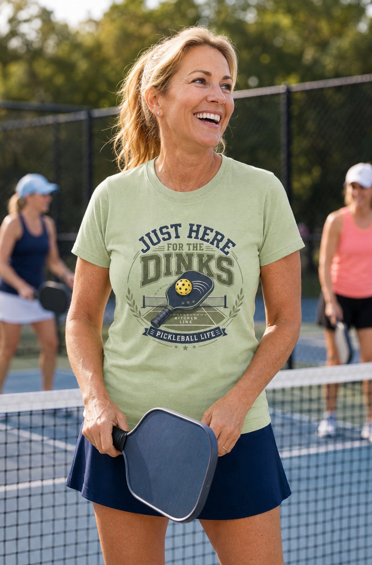 A woman on a pickleball court wearing a pistachio green graphic t-shirt that reads 'JUST HERE FOR THE DINKS' with a pickleball paddle, with other players blurred in the background.