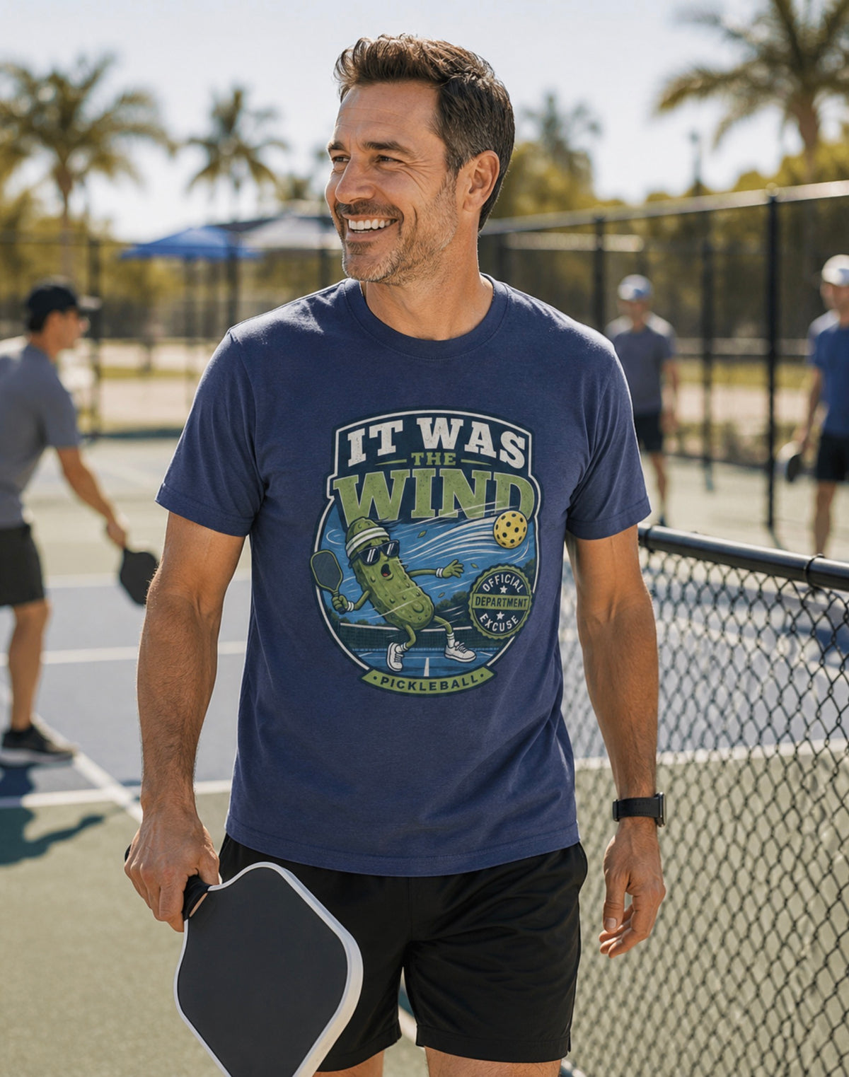 A man on a sunny pickleball court wearing a navy blue crewneck t-shirt featuring a vintage-style circular graphic on the front with a pickleball-themed cartoon character and the text 'IT WAS THE WIND' The Official Excuse Department.