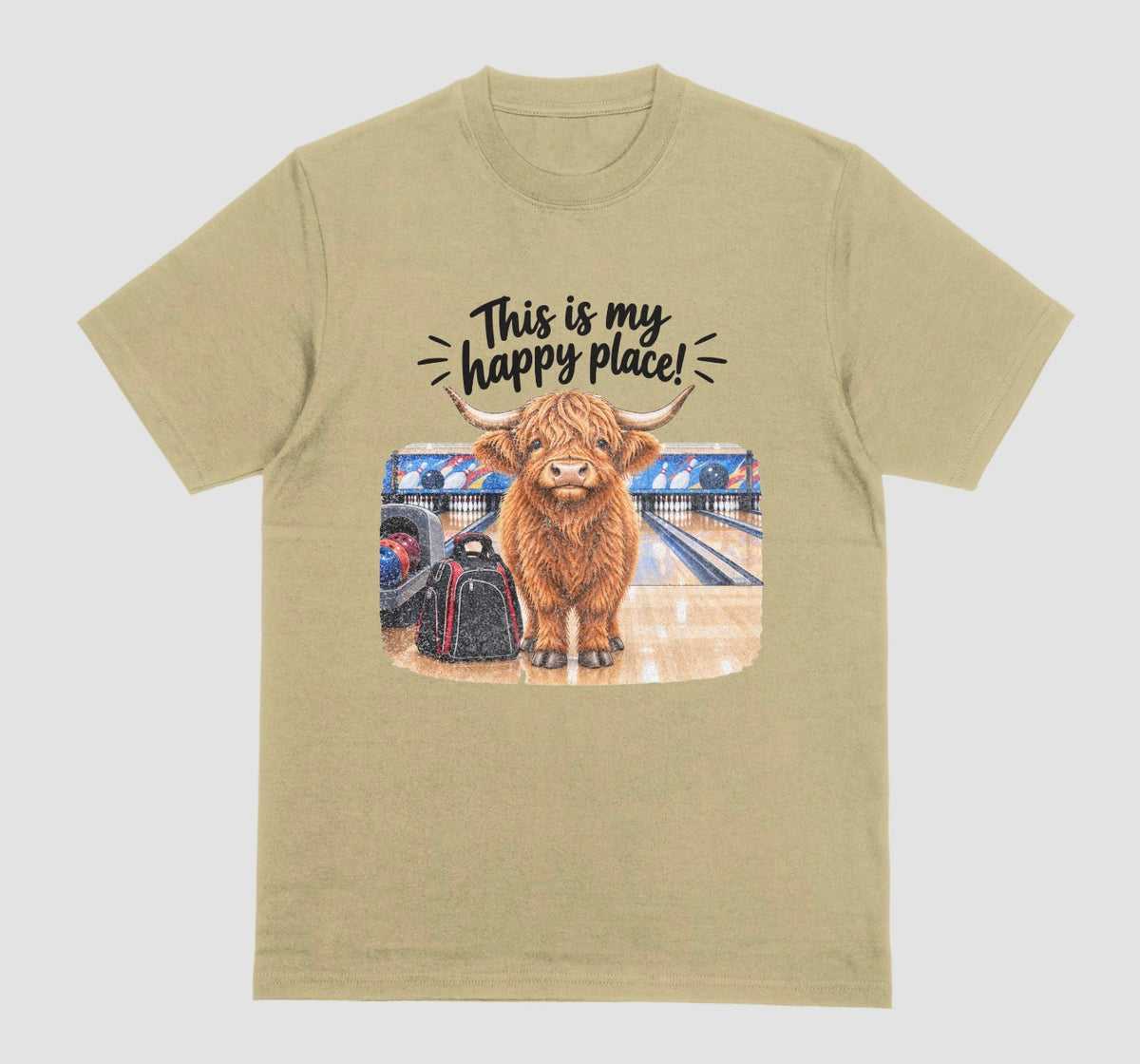 A sand color short-sleeve crewneck t-shirt featuring a graphic of a Highland cow standing on a bowling lane with the text 'This is my happy place!' above it, displayed on a bowling alley background.