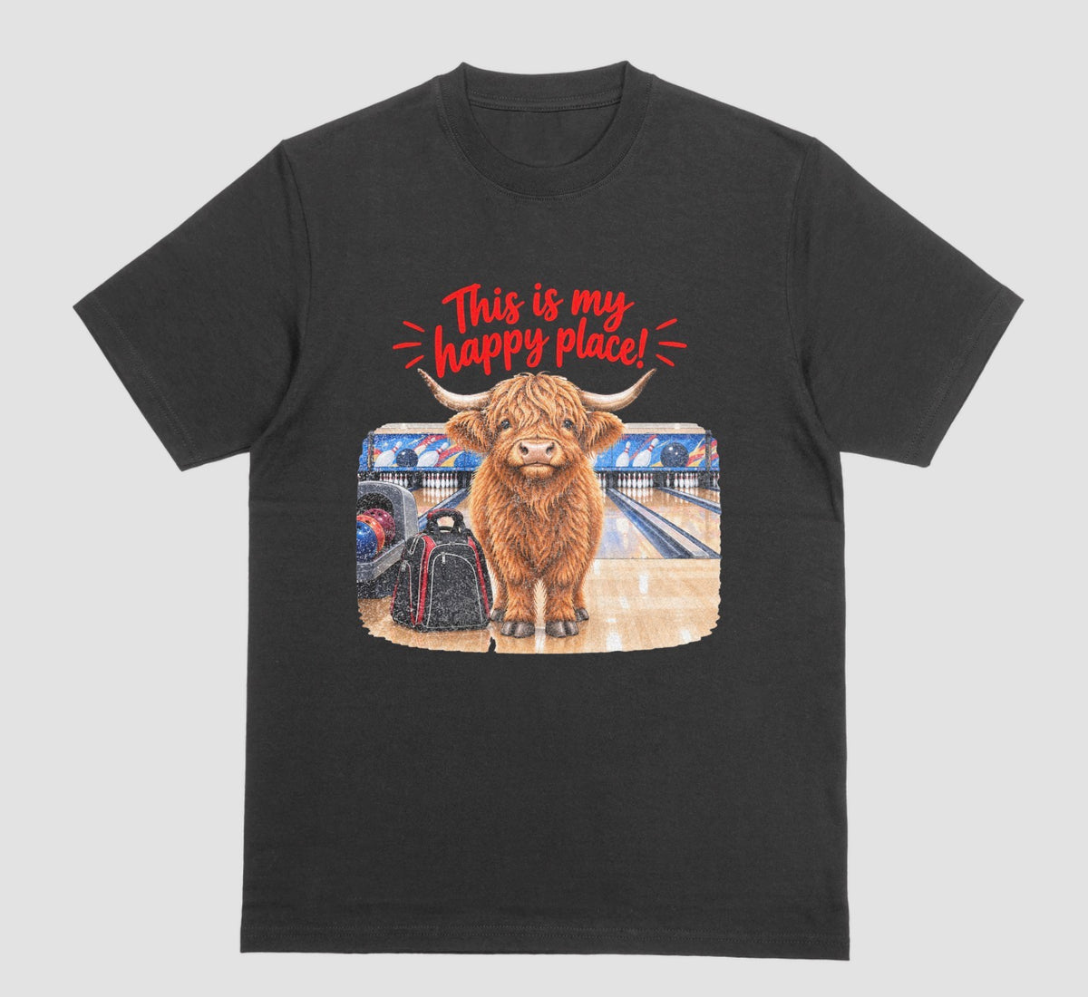 A black short-sleeve crewneck t-shirt featuring a graphic of a Highland cow standing on a bowling lane with the text 'This is my happy place!' above it, displayed on a bowling alley background.