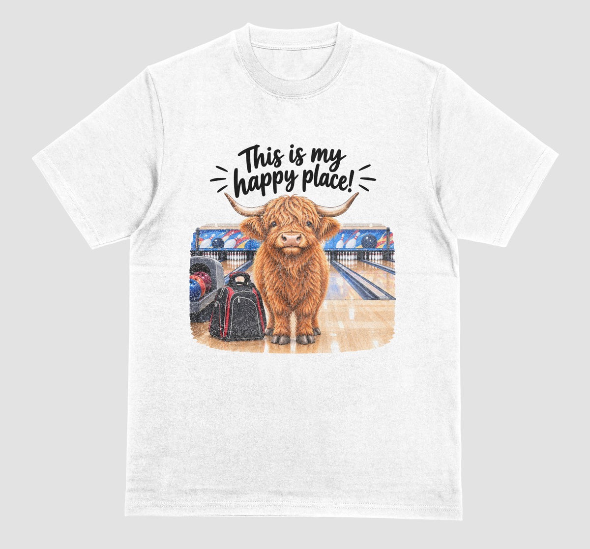 A white short-sleeve crewneck t-shirt featuring a graphic of a Highland cow standing on a bowling lane with the text 'This is my happy place!' above it, displayed on a bowling alley background.