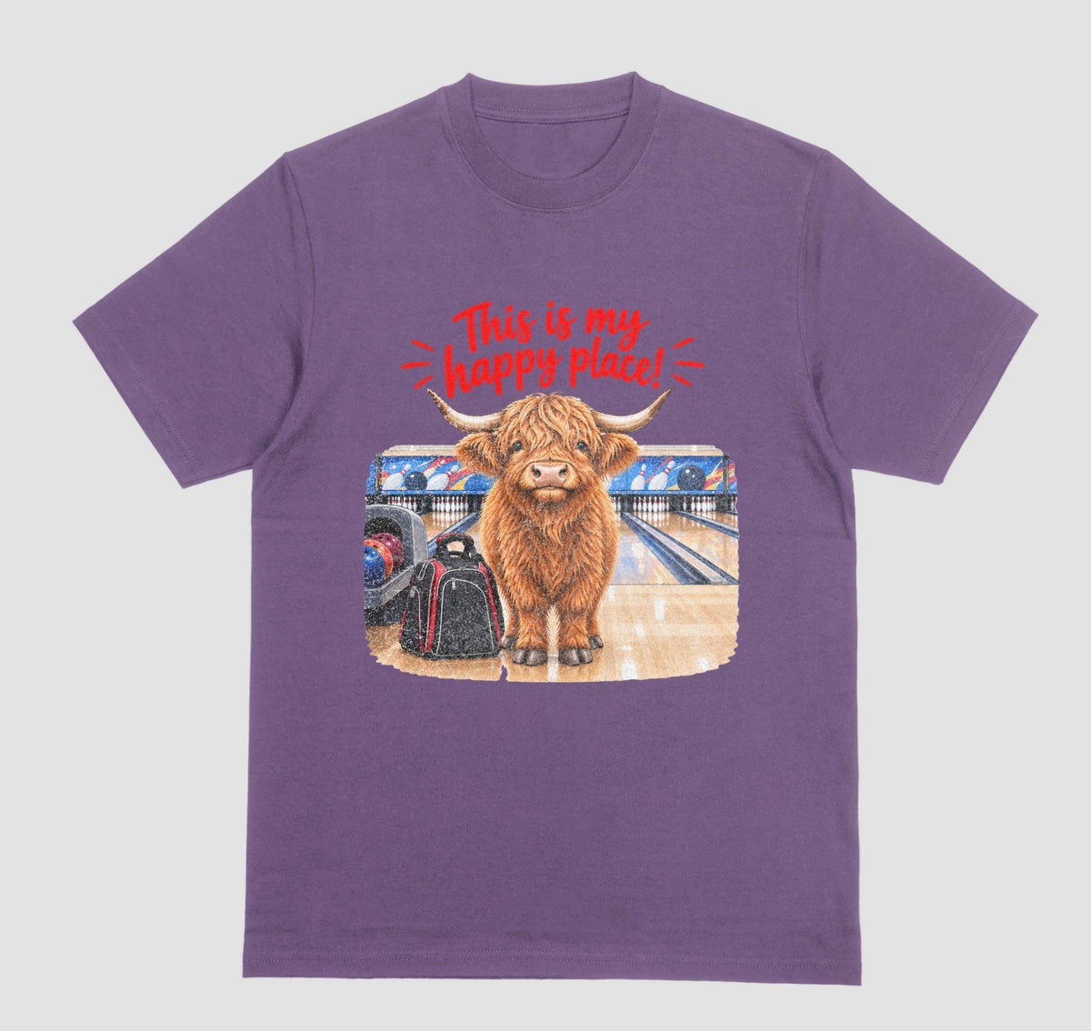 A purple short-sleeve crewneck t-shirt featuring a graphic of a Highland cow standing on a bowling lane with the text 'This is my happy place!' above it, displayed on a bowling alley background.