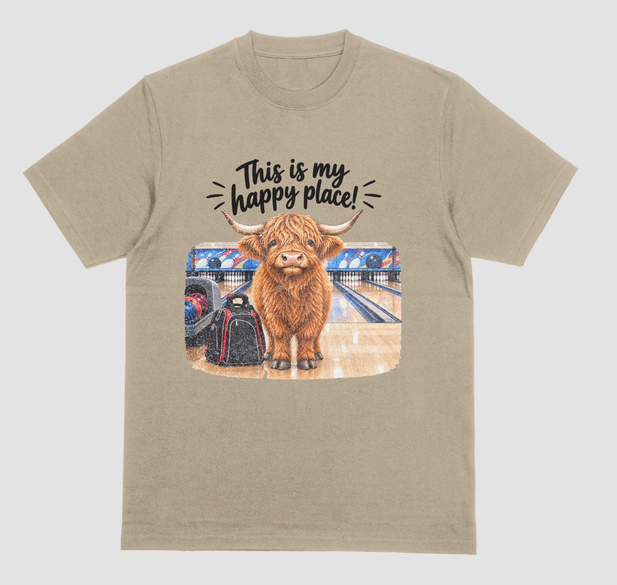 A prairie dust short-sleeve crewneck t-shirt featuring a graphic of a Highland cow standing on a bowling lane with the text 'This is my happy place!' above it, displayed on a bowling alley background.