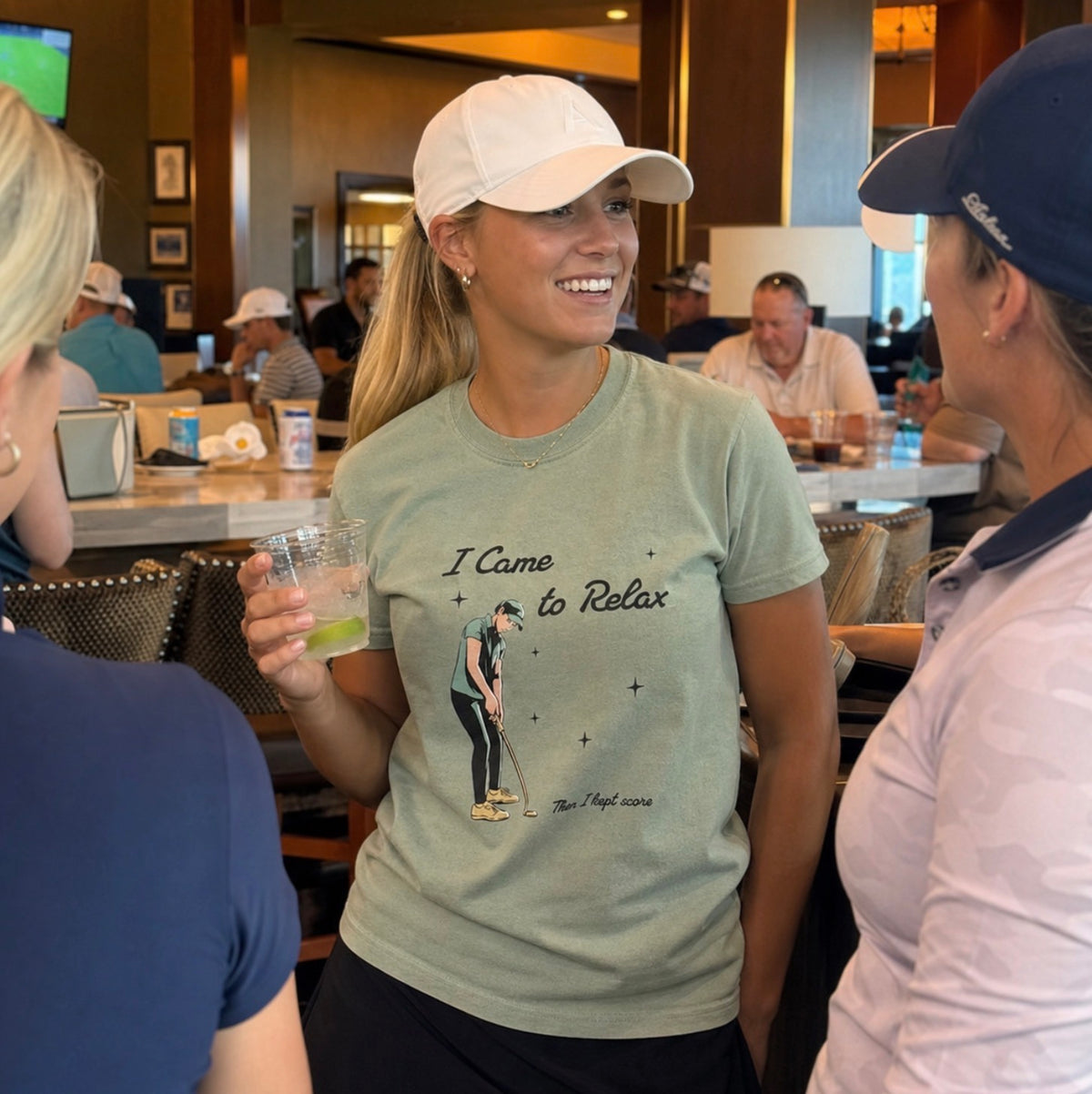A woman in a golf club smiling while wearing a light sage green graphic t-shirt that reads 'I Came to Relax Then I kept score' with a golf club illustration