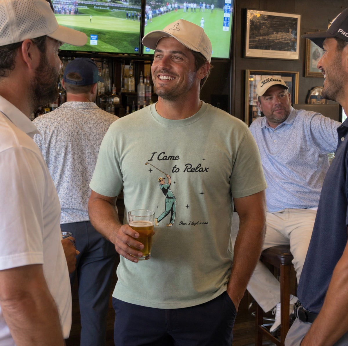 A man in a bar wearing a light sage green graphic t-shirt that reads 'I Came to Relax Then I kept score' with a golfer illustration. He is holding a glass of beer and smiling, with other men and golf game screens visible in the background.