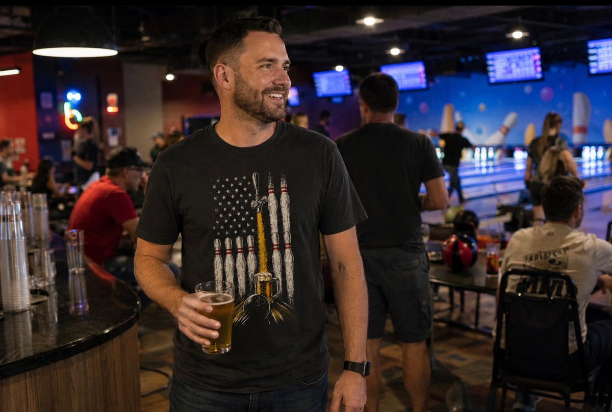 A man in a bowling alley wearing a black graphic t-shirt with a bowling pins and American flag design, a beer tap filling a mug of beer, while a bowling ball travels down the lane and he is holding a pint of beer.