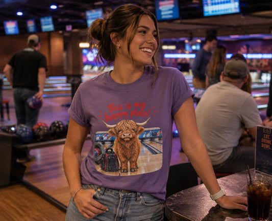 A woman at a bowling alley wearing a purple short-sleeve crewneck t-shirt featuring a graphic of a Highland cow standing on a bowling lane with the text 'This is my happy place!' above it, displayed on a bowling alley background. The background shows bowling lanes and other people in the bowling area.