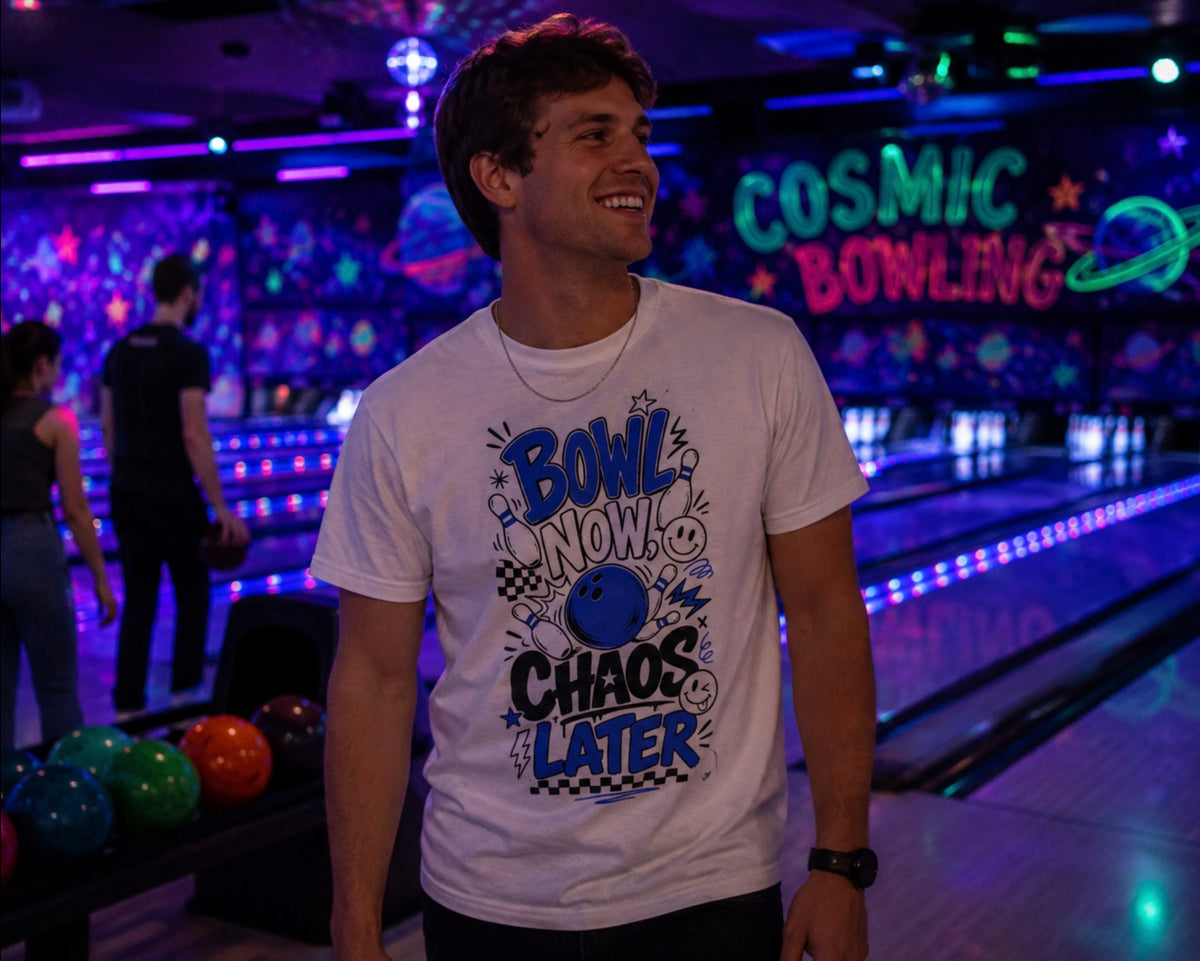 A man smiling in a bowling alley wearing a white graphic t-shirt that reads 'BOWL NOW CHAOS LATER' in blue and black retro-style lettering with a bowling ball illustration. The background features colorful bowling lanes and a neon sign reading 'COSMIC BOWLING'.