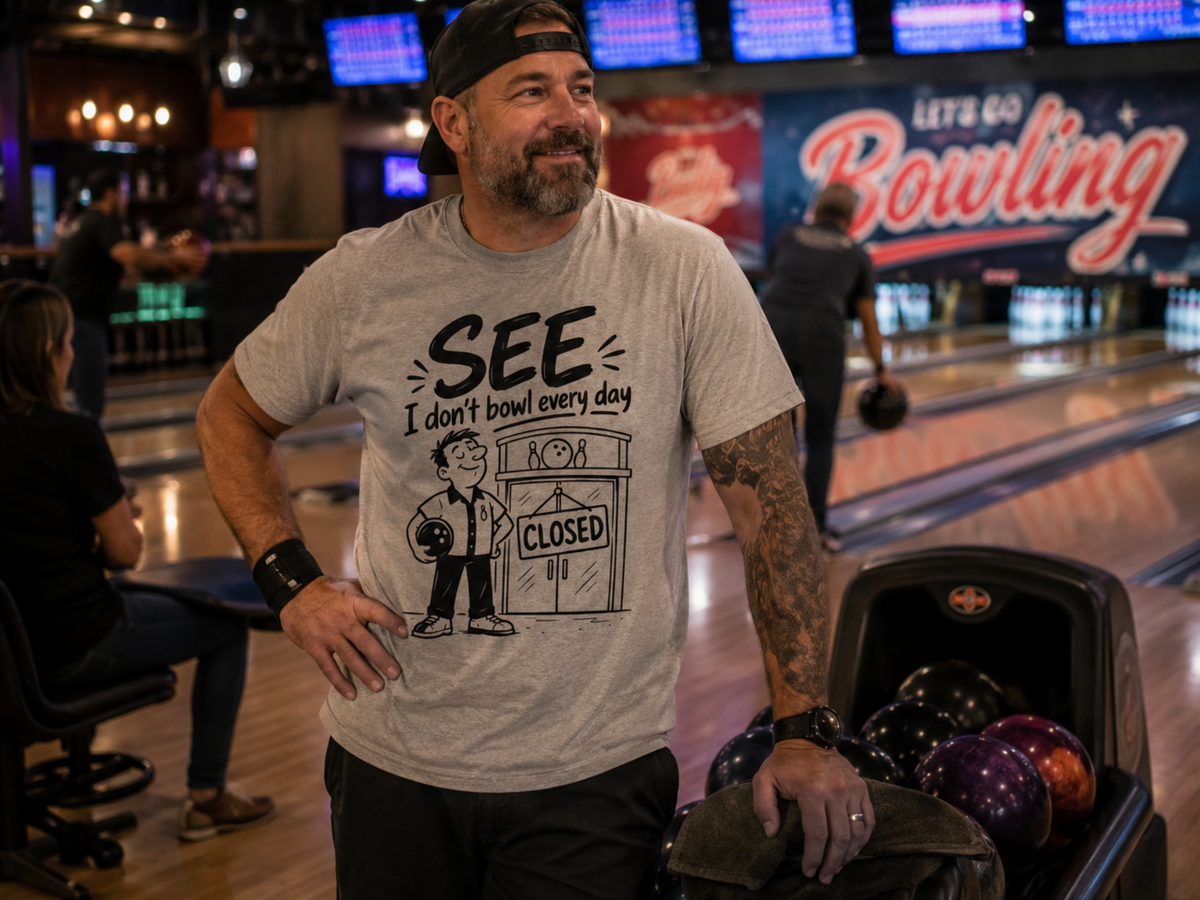 A man in a bowling alley wearing a light gray graphic t-shirt that reads 'SEE I don't bowl every day' with a cartoon bowling pin character and a 'CLOSED' sign. He is standing next to a bowling ball return filled with bowling balls.