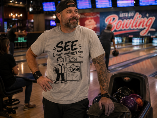 A man in a bowling alley wearing a light gray graphic t-shirt that reads 'SEE I don't bowl every day' with a cartoon bowling pin character and a 'CLOSED' sign. He is standing next to a bowling ball return filled with bowling balls.