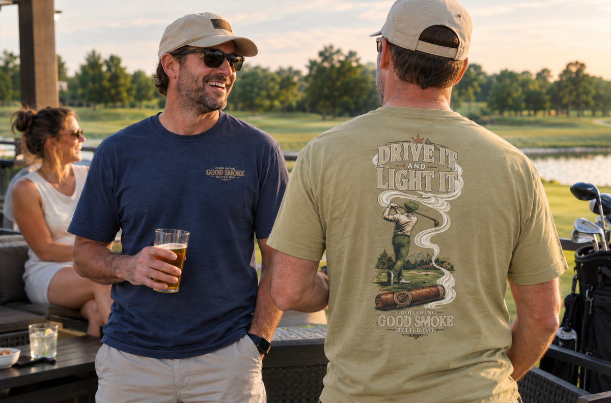 Two men wearing Good Smoke branded t-shirts and caps converse on a patio at sunset, with a golf course and water in the background. One man faces away from the camera, showing the back graphic of his sand colored t-shirt that reads 'DRIVE IT AND LIGHT IT' with a golf ball and smoke illustration.