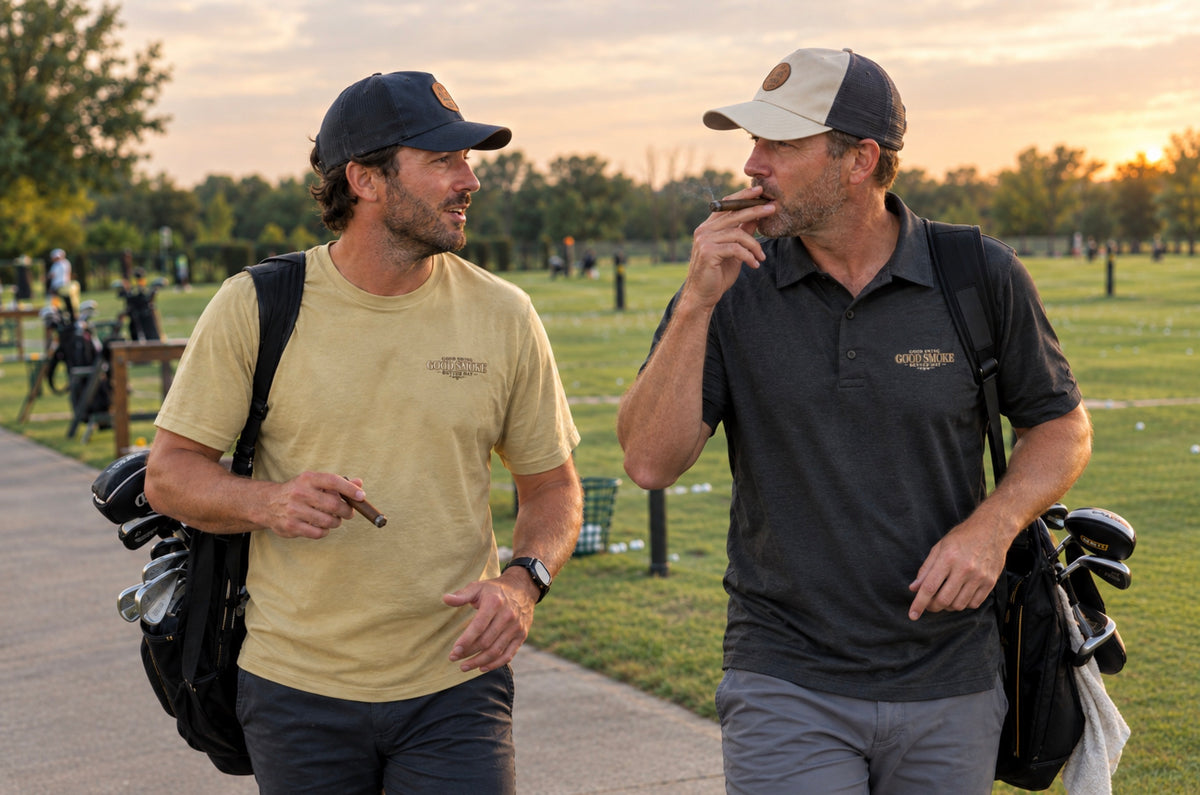 Two men walking and talking on a golf course path at sunset, one wearing a sand color t-shirt Front chest features small text reading 'Good Swing, Good Smoke, Better Day.', and the other wearing a black polo shirt Front chest features small text reading 'Good Swing, Good Smoke, Better Day.'  