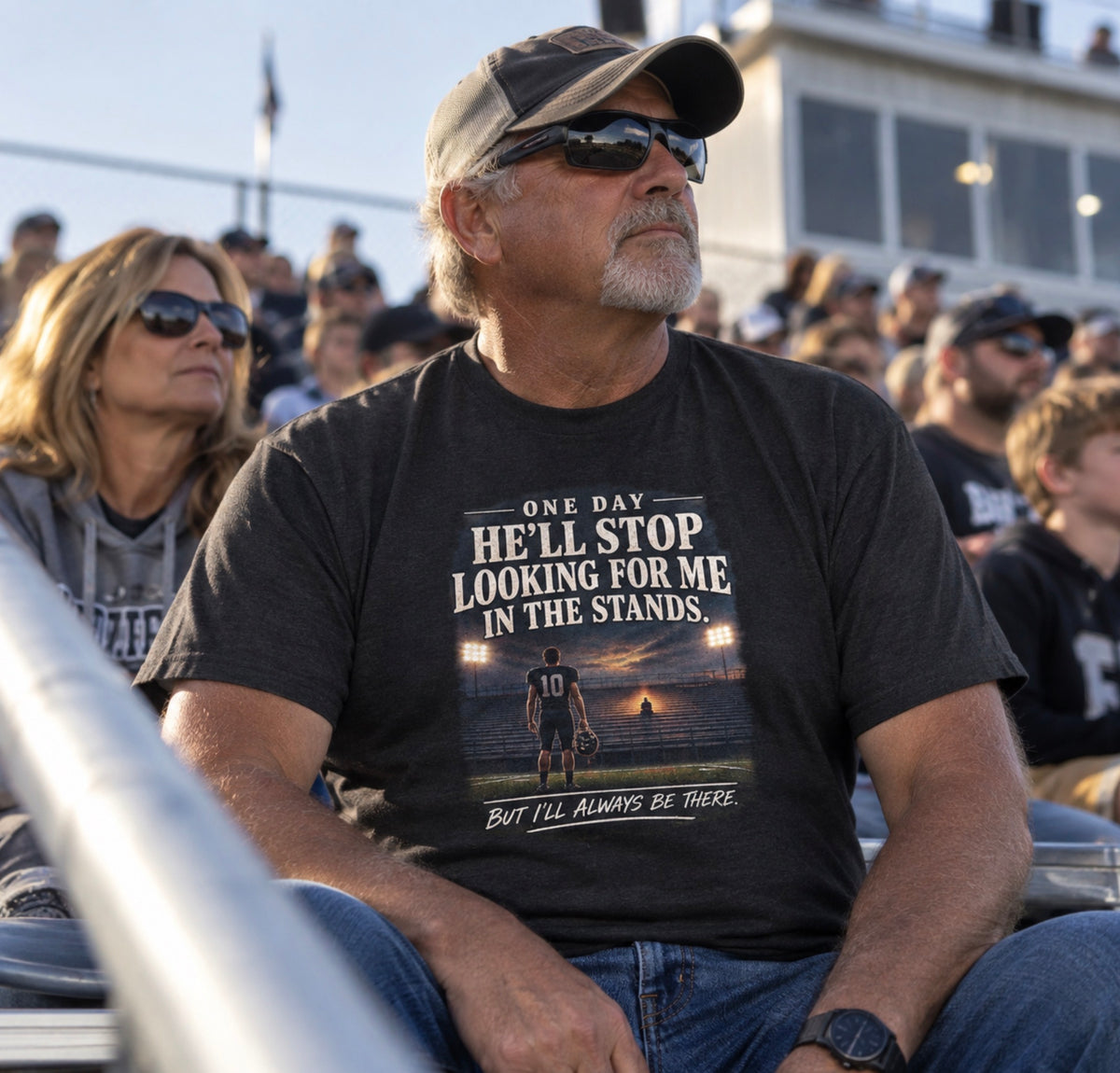 A man wearing a black graphic t-shirt and sunglasses sits in a crowded outdoor stadium. The t-shirt features a printed graphic of a football player on a beach at sunset with the text 'ONE DAY HE'LL STOP LOOKING FOR ME IN THE STANDS.
