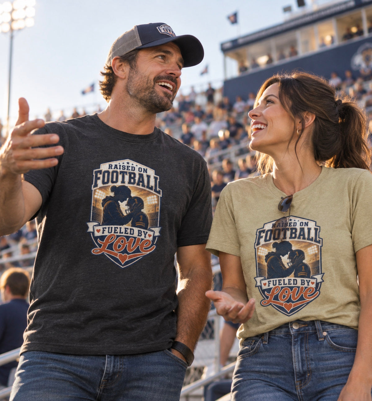 A man and a woman standing in a stadium, both wearing matching graphic T-shirts that read 'Raised on Football Fueled by Love' with a football player graphic. The man wears a black T-shirt and cap, and the woman wears a sand color T-shirt and jeans.
