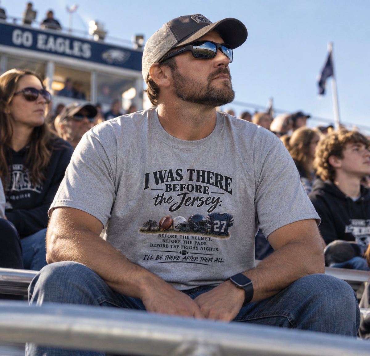 A man sitting in stadium seating at a football game, wearing a light gray graphic t-shirt that reads 'I WAS THERE BEFORE the Jersey'.