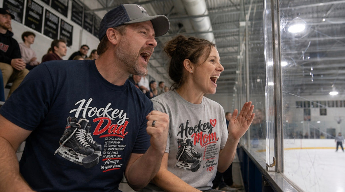Man in navy Hockey Dad t-shirt and woman in grey Hockey Mom t-shirt cheering at a hockey game from behind the glass