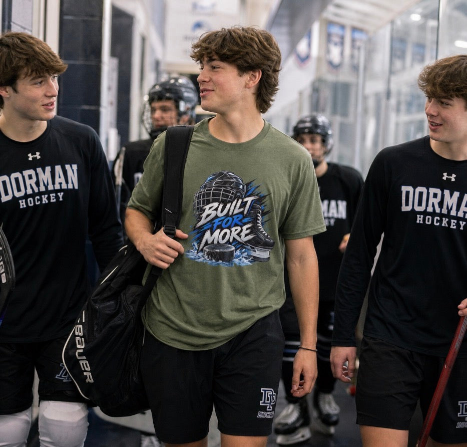 Teen hockey player in an olive green "Built For More" t-shirt carrying a gear bag in a rink hallway with teammates