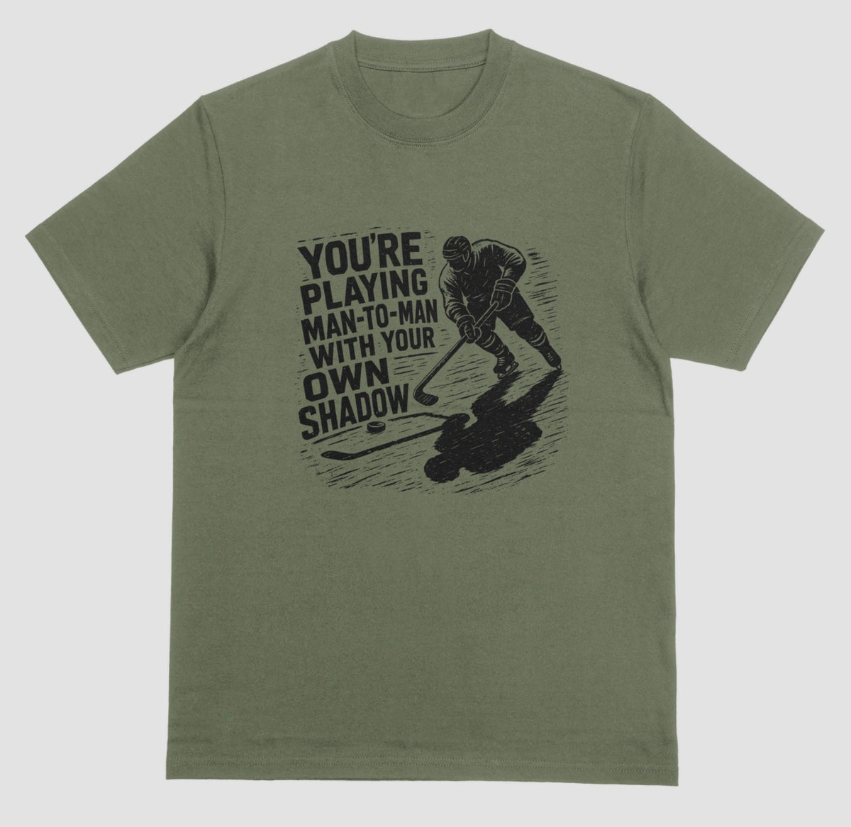 A military green hockey t-shirt with black graphic of a skater and his long shadow, reading “You’re playing man-to-man with your own shadow”