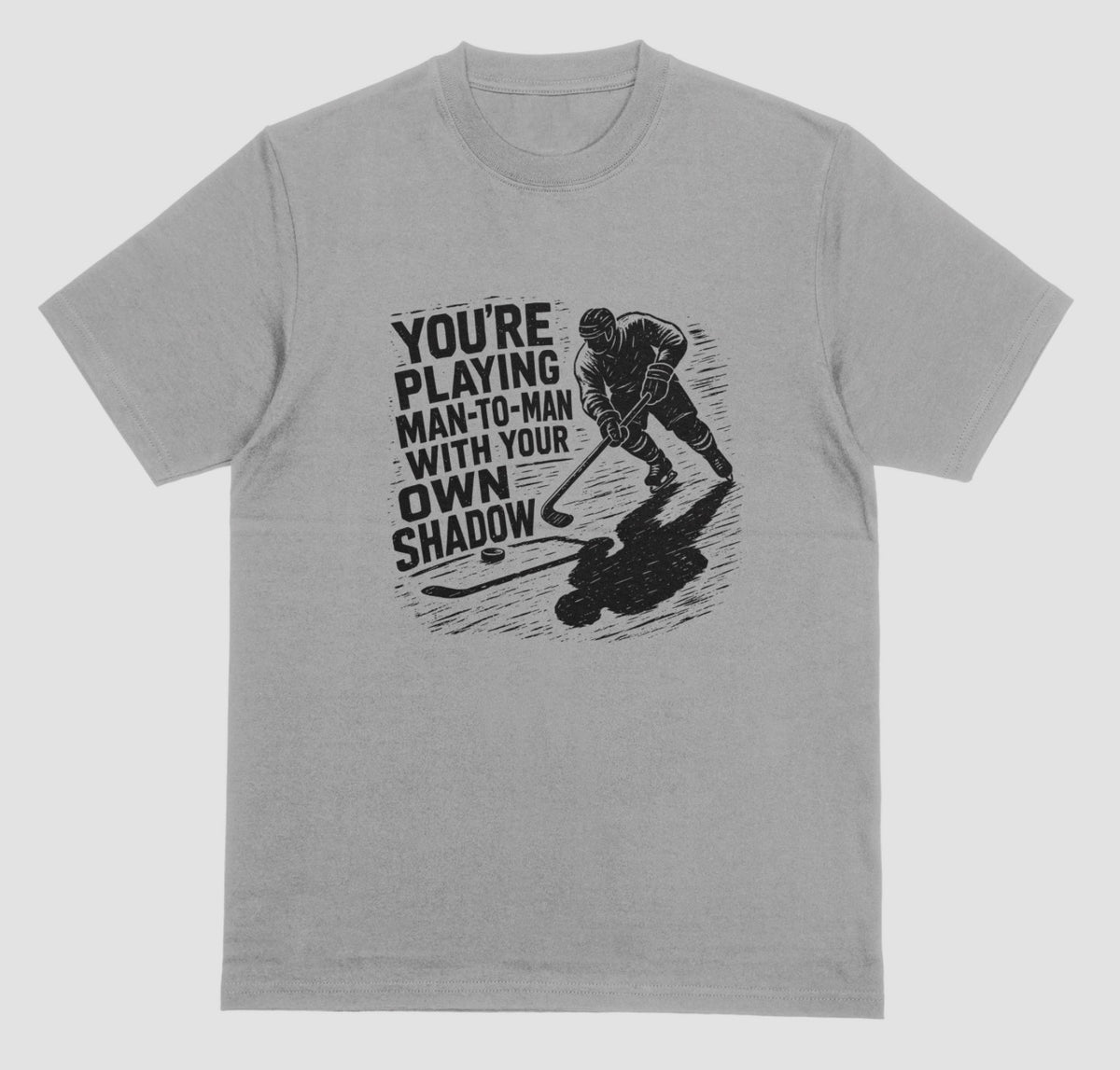 A grey hockey t-shirt with black graphic of a skater and his long shadow, reading “You’re playing man-to-man with your own shadow”