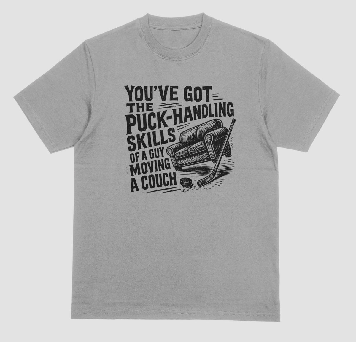 A grey hockey humor t-shirt with black graphic of a couch, hockey stick, and puck, reading “You’ve got the puck-handling skills of a guy moving a couch”