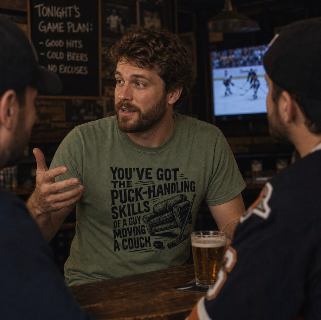 Bearded man wearing olive green t-shirt reading “You’ve got the puck-handling skills of a guy moving a couch” with couch and hockey stick graphic, talking with friends at a bar