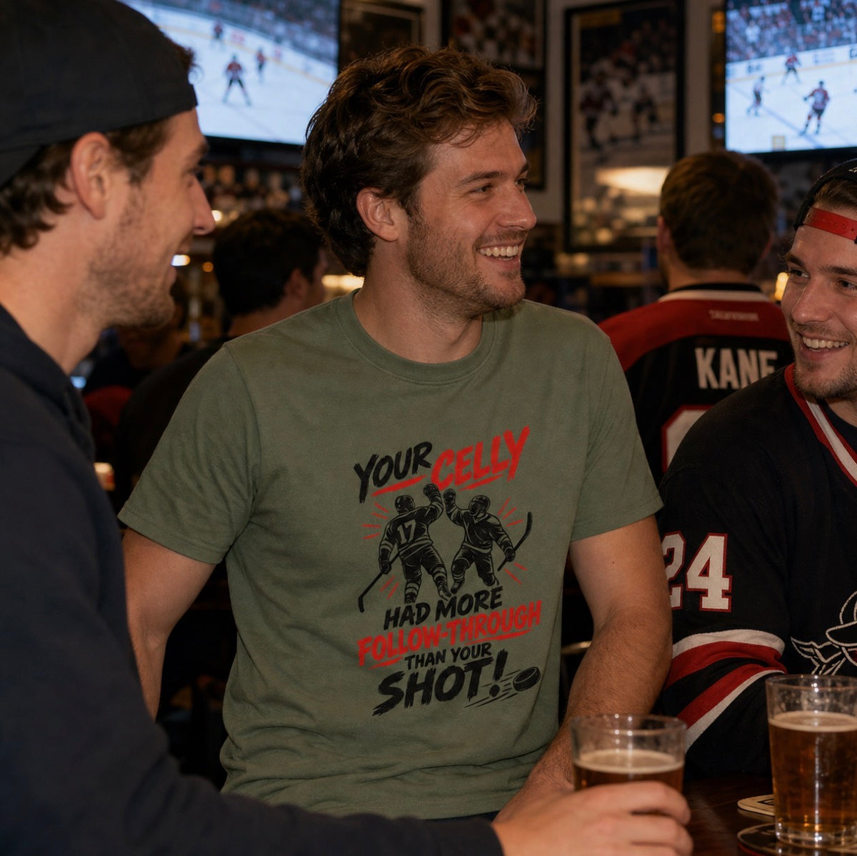Smiling man wearing olive green hockey t-shirt with red and black graphic reading “Your celly had more follow-through than your shot,” at a sports bar with friends
