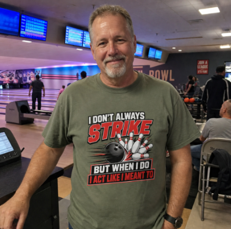 A man standing in a bowling alley wearing a graphic t-shirt that reads 'I DON'T ALWAYS STRIKE BUT WHEN I DO I ACT LIKE I MEANT TO' with a bowling ball and pins design.
