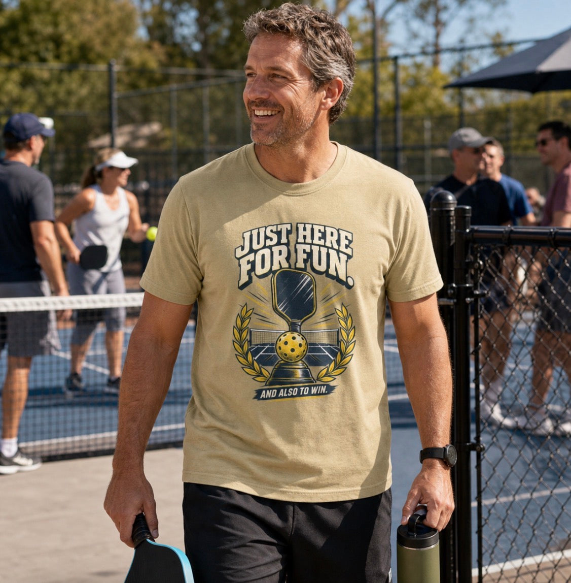 A man smiling and wearing a Funny pickleball shirt with a trophy, paddle and laurel wreath design that says Just Here for Fun and Also to Win standing on a pickleball court