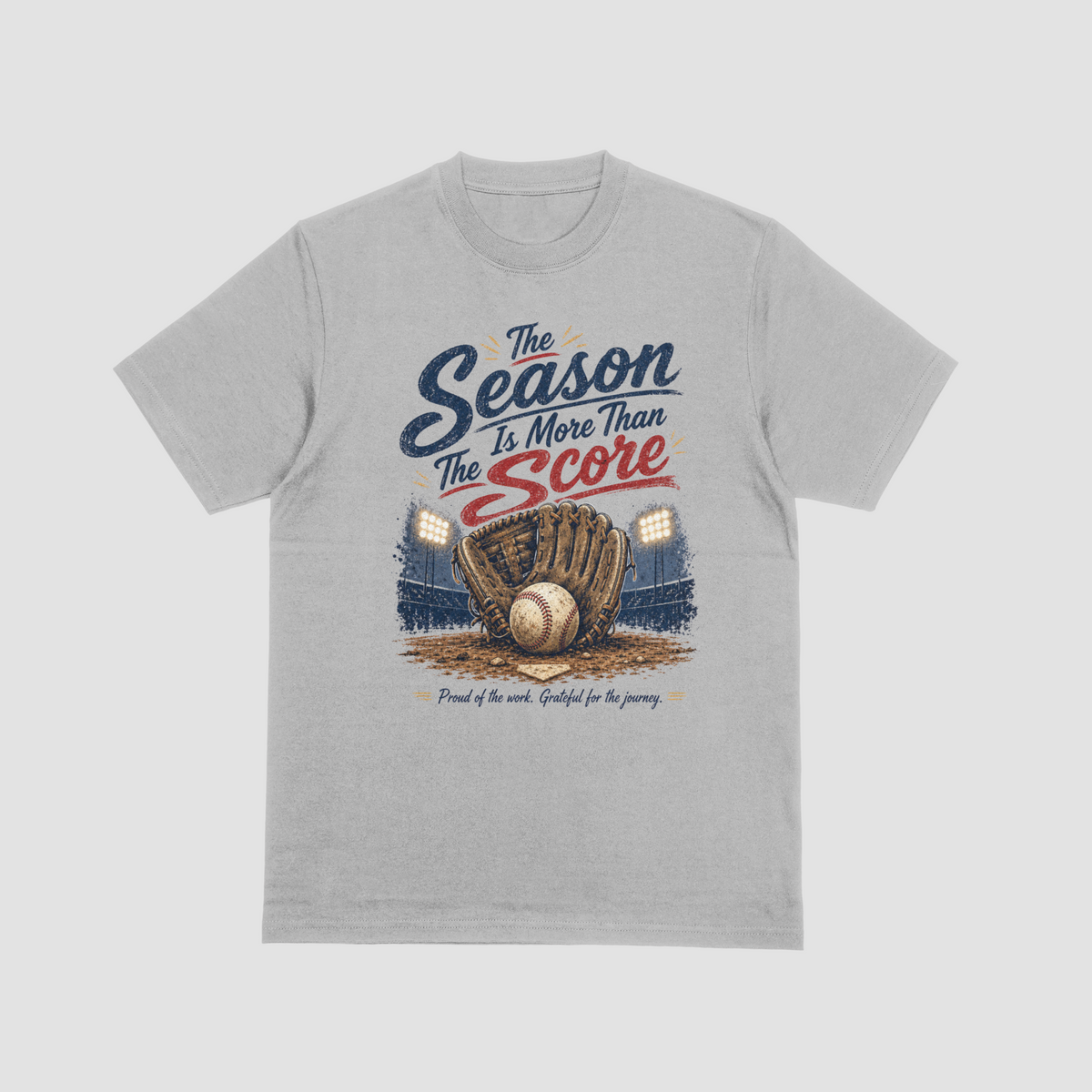 A grey short-sleeve crewneck t-shirt featuring a vintage-style baseball graphic. The design reads “The Season Is More Than The Score” in navy blue and red lettering above a brown leather baseball glove holding a baseball, with a baseball field and illuminated stadium lights in the background. The design also includes the phrase “Proud of the work. Grateful for the journey.”