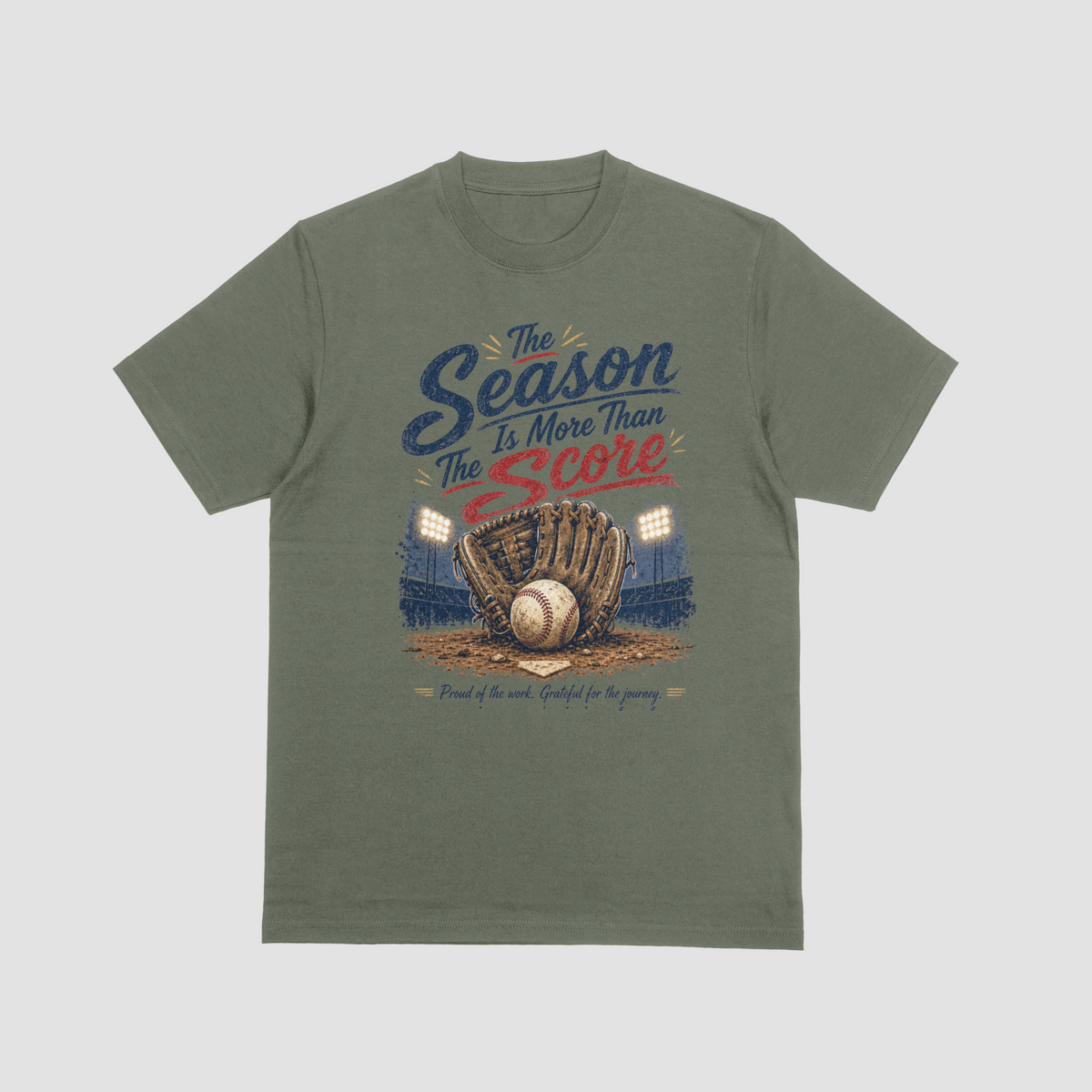 A military green short-sleeve crewneck t-shirt featuring a vintage-style baseball graphic. The design reads “The Season Is More Than The Score” in navy blue and red lettering above a brown leather baseball glove holding a baseball, with a baseball field and illuminated stadium lights in the background. The design also includes the phrase “Proud of the work. Grateful for the journey.”