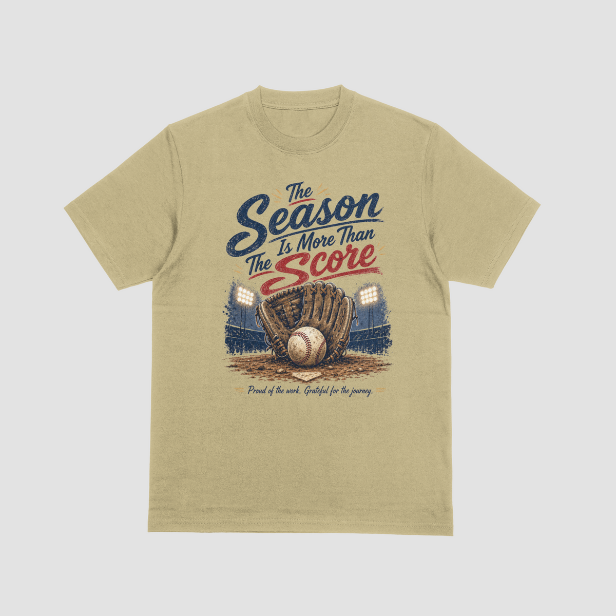 A sand color short-sleeve crewneck t-shirt featuring a vintage-style baseball graphic. The design reads “The Season Is More Than The Score” in navy blue and red lettering above a brown leather baseball glove holding a baseball, with a baseball field and illuminated stadium lights in the background. The design also includes the phrase “Proud of the work. Grateful for the journey.”